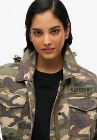 Veste camouflée en vert, marron et beige avec une fermeture éclair sur le devant, un grand col et des patches. Portée par-dessus un haut noir, avec une étiquette de marque sur la poitrine.