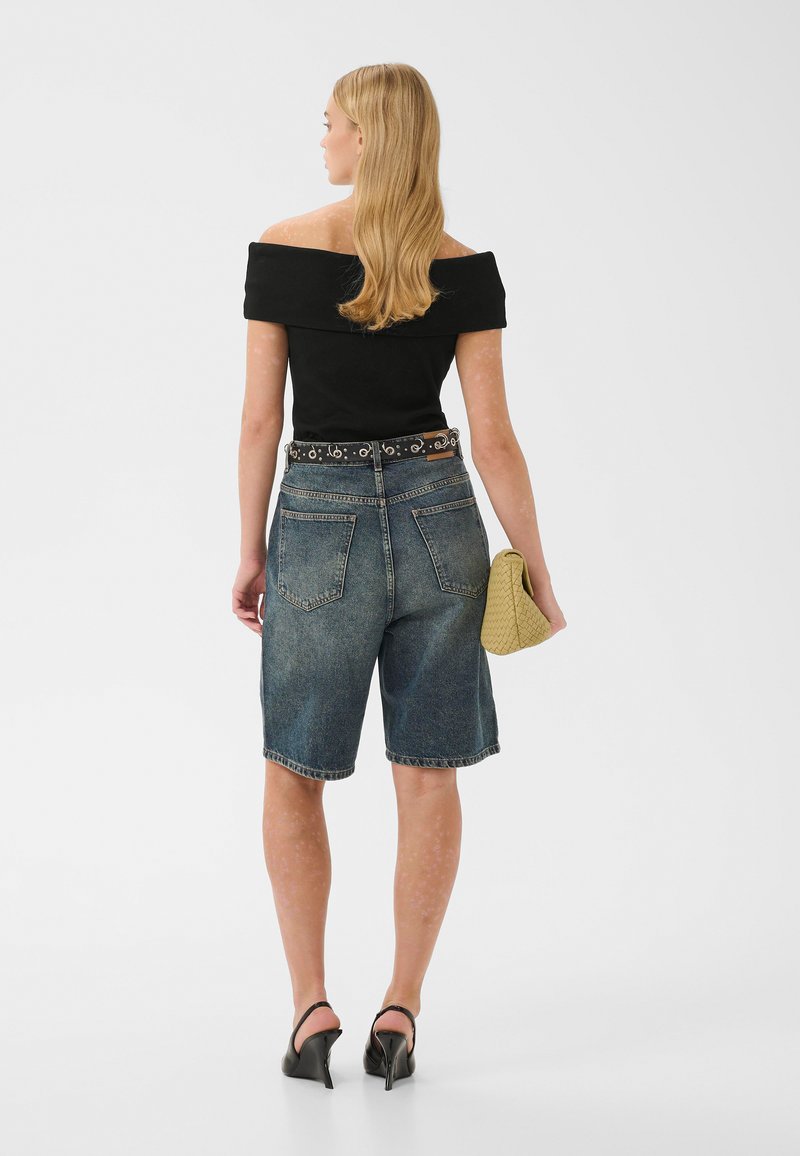Femme aux longs cheveux blonds portant un top noir épaules dénudées, un short en jean taille haute, des talons noirs, tenant une pochette tissée jaune, debout, dos tourné.