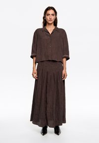 Brązowy, lniany zestaw dwuczęściowy, składający się z zapinanej na guziki bluzki z krótkimi rękawami oraz długiej plisowanej spódnicy. Całość dopełniają czarne, spiczaste buty.