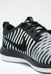 Svart- och vitmönstrad stickad träningissko med texturerat randigt mönster, rundad tå, snörning och svart gummisula.