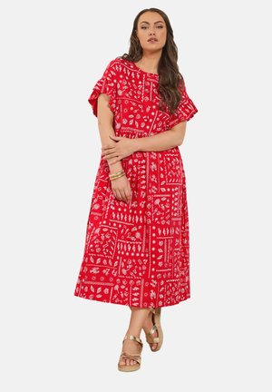 Kvinde iført rød midi-kjole med hvide blomster- og abstrakte mønstre, flæseærmer, gyldne sandaler og gyldne armbånd, stående med armene over kors.