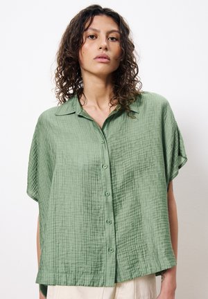 Chemise courte à manches courtes de couleur verte texturée avec un col, comportant une fermeture à boutons et une coupe ample. Le tissu a un aspect froissé.