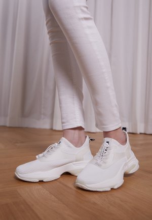Osoba nosząca białe wąskie spodnie i białe masywne sneakersy na drewnianej podłodze z jasnymi zasłonami w tle.