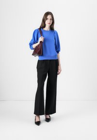 Felpa blu con maniche a sbuffo, abbinata a pantaloni ampi neri e décolleté neri a punta. Il modello tiene una borsa rosso scuro.