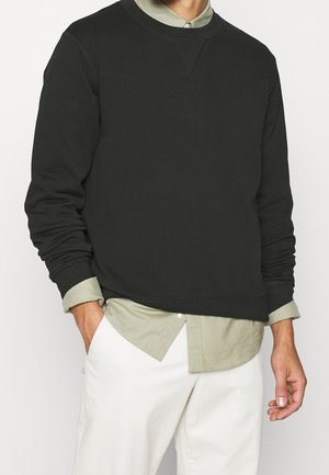 Felpa nera con colletto e polsini a coste, che presenta una vestibilità comoda su una maglietta verde oliva. I pantaloni bianchi completano l'outfit.