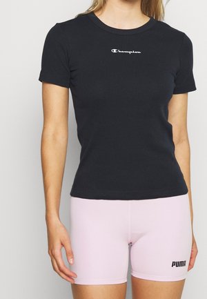 Kobieta w dopasowanym czarnym t-shircie Champion i jasnoróżowych szortach rowerowych Puma, stojąca na tle jednolitego jasnego tła.