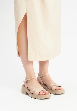 Beige platform-sandaalit ristikkäisillä hihnoilla, nilkkahihna soljella, teksturoitu pohja ja jalkatuki, joka paljastaa varpaat.