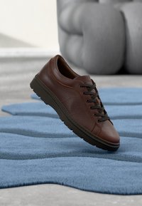 Baskets en cuir marron avec finition texturée, bout rond et lacets foncés. Semelle en caoutchouc à profil bas. Placées sur un tapis bleu.