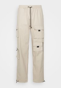 Beige cargobukser med elastisk talje, sort snor, flere klaplommer og lynlåse detaljer på en ens hvid baggrund.