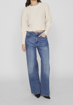 Pull court en maille crème associé à un jean large taille haute bleu clair et des talons noirs à bout pointu. Maille texturée et tissu en denim.