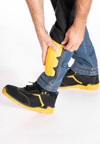 Bottes de sécurité noires et jaunes avec une tige texturée et une semelle en caoutchouc. Une protection rembourrée jaune est ajustée sur une jambe vêtue de denim.