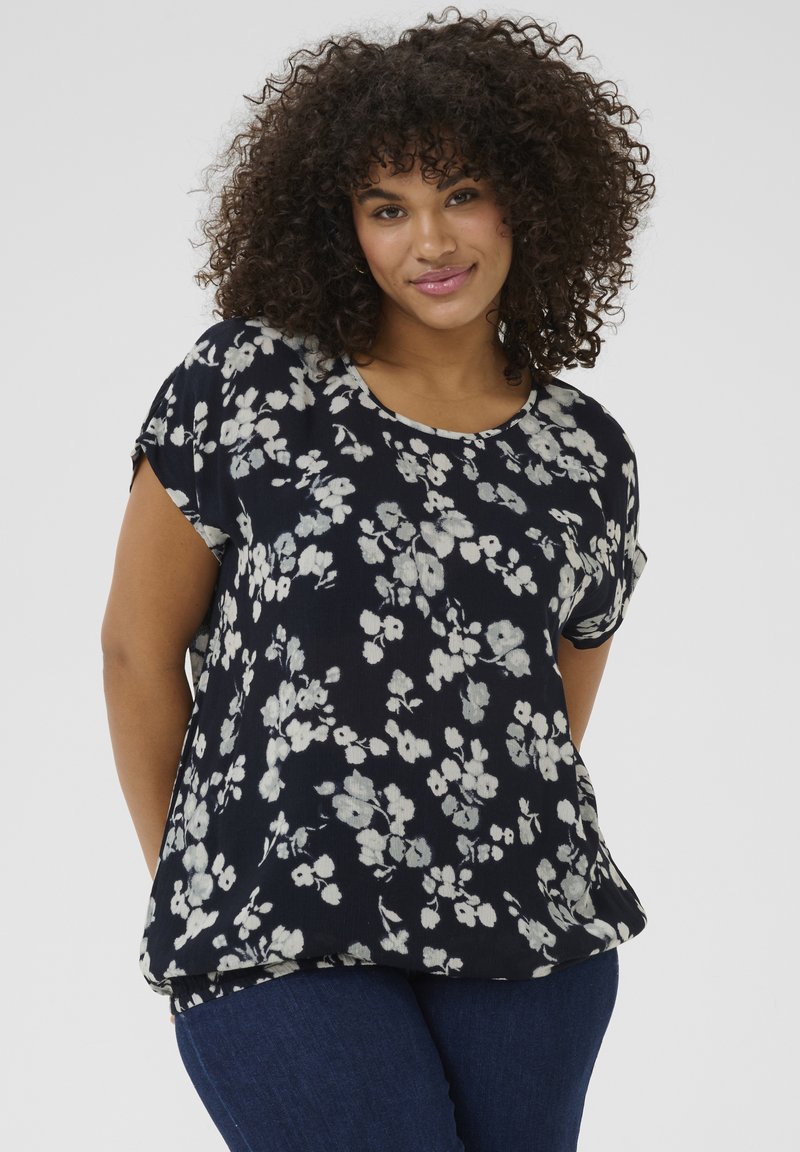 Blusa blu navy con motivo floreale bianco, maniche corte, scollatura rotonda e vestibilità ampia, abbinata a jeans scuri. Tessuto morbido e leggero.