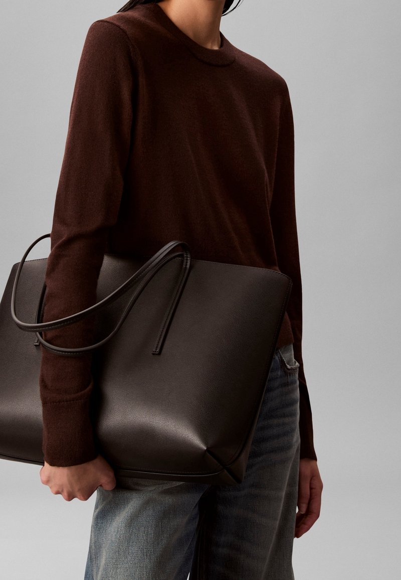 Personne portant un pull marron foncé et un jean bleu tenant un grand sac cabas en cuir marron foncé sur un fond gris.