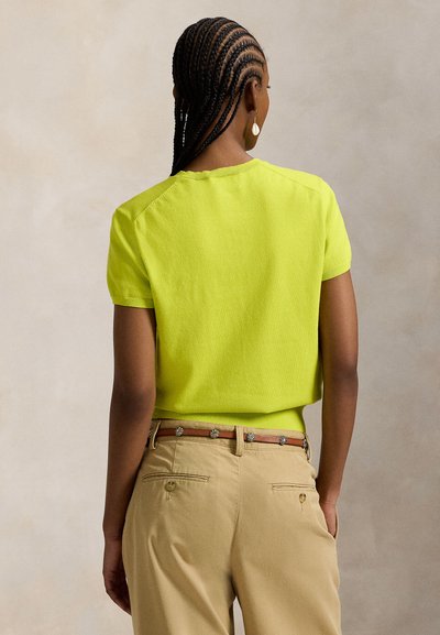 Κοντομάνικο, φωτεινό κίτρινο τοπ με στρογγυλή λαιμόκοψη και υφή, συνδυασμένο με ανοιχτό χακί παντελόνι και διακοσμητική ζώνη.