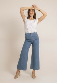 Pantalon en denim taille haute, large, avec des boutons sur le côté, associé à un haut blanc uni à manches courtes et des escarpins dorés à lanières.