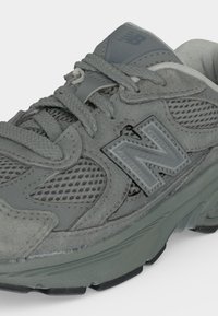 Γκρι αθλητικό παπούτσι New Balance με πάνελ από δίχτυ και σουέντ, παχιά σόλα, δεμένα κορδόνια και έντονο λογότυπο "N" στο πλάι.