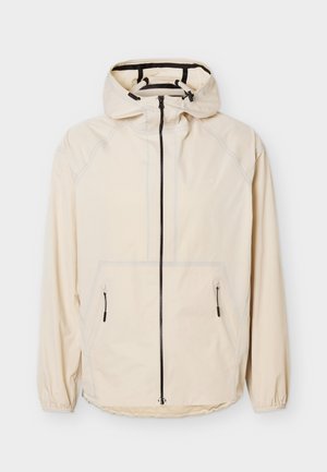 Svetlo bež jakna s kapuco, sprednjo zadrgo, dvema žepoma z zadrgo in elastičnimi zapestji, položena na ravno belo podlago.