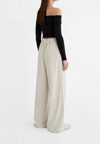 Zwart langarm off-the-shoulder top gecombineerd met losse crèmewitte broek met een elastische tailleband. Gladde stoftextuur.