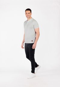 Jasnoszara koszulka polo z krótkim rękawem, z kołnierzykiem i brązową skórzaną metką na dolnym brzegu, w połączeniu z czarnymi dżinsami slim fit i białymi trampkami.