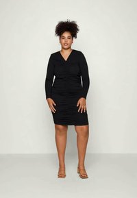 Mulher com cabelo encaracolado, vestindo um vestido preto ajustado de manga longa e sandálias de salto em tom claro, de pé contra um fundo claro e liso.