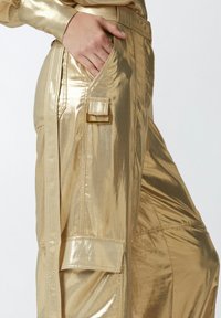 Zlatne metalik cargo hlače s sjajnim završetkom, s bočnim džepom s preklopom, petljama za pojas i opuštenim krojem. Glatka tekstura kroz cijele hlače.