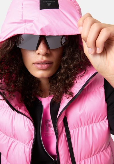 Różowa kamizelka puchowa z kapturem, z czarnymi akcentami. Nosi ją nad czarną koszulą i różową bluzką. Modelka ma na sobie ciemne okulary przeciwsłoneczne.