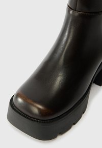 Καφέ δερμάτινη μπότες μέχρι τον αστράγαλο με στρογγυλή μύτη, υφασμάτινη υφή, παχύ μαύρο πάτο και ορατές ραφές κατά μήκος των άκρων.