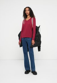 Bordeaux V-hals trui met witte en rode strepen, gecombineerd met uitlopende blauwe jeans en zwarte platform schoenen, terwijl ze een zwarte gewatteerde jas vasthoudt.