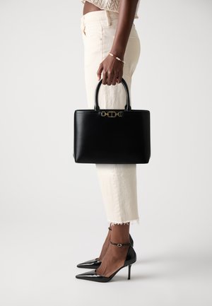 Black leather handbag with structured design, gold hardware, and dual handles. Paired with cream-colored pants and black pointed-toe heels.