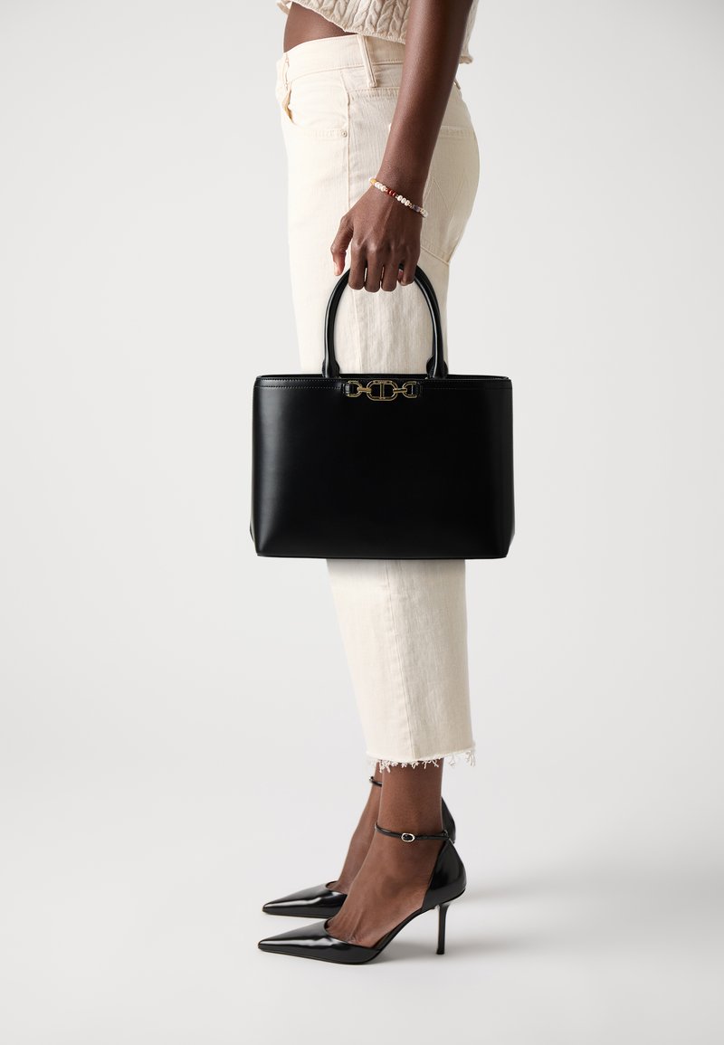 Black leather handbag with structured design, gold hardware, and dual handles. Paired with cream-colored pants and black pointed-toe heels.