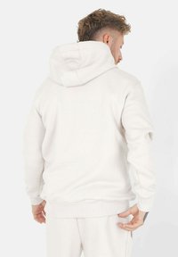 Hvit hettegensser laget av myk stoff, med en stor hette, ribbede mansjetter og en avslappet passform. Sømdetaljer er synlige på baksiden.