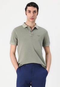Polo de culoare verde măsliniu dintr-un amestec de bumbac, cu guler cu două butoane, mâneci scurte și logo brodat. Asortat cu pantaloni bleumarin.