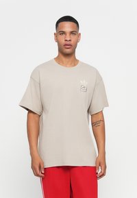 Beige bomulds T-shirt med korte ærmer, rund hals og et lille grafisk logo på venstre bryst. Glat tekstur, afslappet pasform.