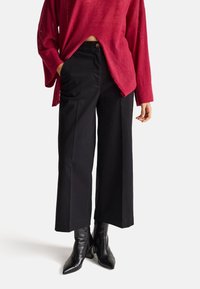 Černé široké kalhoty s předními kapsami, spárovány s volným červeným topem. Součástí oblečení jsou lesklé černé kotníkové boty s krokodýlí texturou.