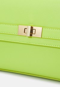 Limoengroene handtas met een gestructureerde synthetische buitenkant, een gladde sluiting van goudkleurig metaal en decoratief stiksel langs de naden.