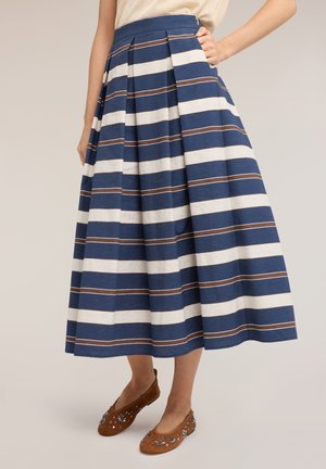 Kobieta w plisowanej spódnicy midi z poziomymi niebieskimi, białymi i brązowymi paskami, w zestawieniu z brązowymi zdobionymi płaskimi butami.