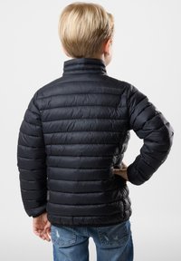 Sort pufferjakke med horisontalt quiltet design, høj krave og tekstureret overflade. Kombination med blå slidte jeans.
