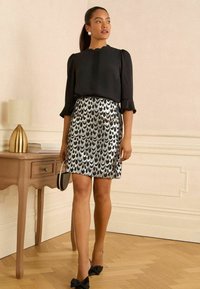 Blusa negra con escote en forma de pico y mangas con volantes, combinada con una falda plateada cubierta de lentejuelas que presenta un patrón de olas. Tacones adornados con un lazo negro.