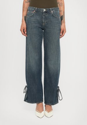 Jeans a gamba larga di media lavatura con lacci alla caviglia indossati da una persona tatuata con décolleté bianchi a punta, mostrati dalla vita ai piedi.