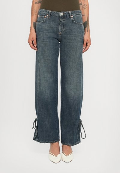 Mid-wash wide-leg jeans with ankle ties worn by a tattooed person in white pointed-toe heels, shown from waist to feet.