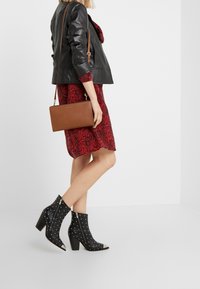 Schwarze Lederstiefeletten mit silbernen Nieten und spitzen Zehen, kombiniert mit einem rot gemusterten Kleid und einer braunen Umhängetasche.