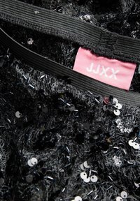 Černý flitrovaný top s průhlednou texturou, obsahující růžovou cedulku s nápisem "JJXX." Zahrnuje černé elastické ramínka a různé velikosti a tvary flitrů.