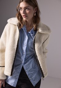 Junge Frau trägt eine cremefarbene Sherpa-Jacke über einem blau-weiß gestreiften Hemd mit Knöpfen, schaut zur Seite und trägt goldene Kreolen.