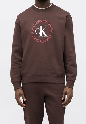 Mężczyzna noszący brązową bluzę i dopasowane spodnie, bluza ma duże białe logo "cK" z czerwonym okrągłym napisem "Calvin Klein New York City Since 1968".