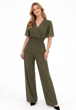 Kobieta ubrana w oliwkowozielony kombinezon z szerokimi nogawkami, z dekoltem w serek, elastyczną talią, falbanowymi rękawami i czarnymi szpilkami, stojąca na białym tle.