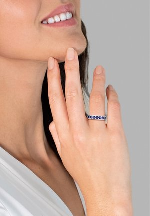 Ringen har et sølvbånd prydet med blå ædelstene, båret på en hånd, der blidt rører ved hagen, hudtonen er lys, baggrunden er grå.