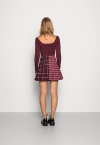 Tmavě červený top s dlouhým rukávem a širokým výstřihem, kombinovaný s plisovanou sukní s dvěma károvanými vzory v odstínech bordó a růžové. Černé boty.