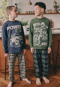 Due ragazzi indossano pigiami a manica lunga: uno blu navy con una grafica di veicolo, l'altro verde con una grafica di motocicletta. Entrambi indossano pantaloni a quadri.
