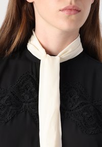 Schwarze Bluse mit filigranen schwarzen Spitzenbesätzen am Ausschnitt, versehen mit einem weichen elfenbeinfarbenen Band, das um den Kragen gebunden ist, um zusätzliche Eleganz zu verleihen.