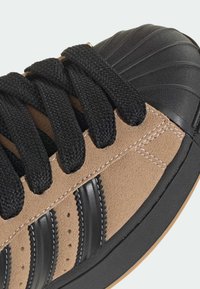 Semišová teniska svetlohnedej farby s čiernymi gumovými detailmi, vybavená textúrovanými čiernymi šnúrkami a tromi čiernymi pruhmi na boku.